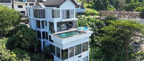 A modern multi-story building with a rooftop pool, surrounded by lush greenery and other residential structures.