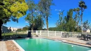 A swimming pool with a stone border, surrounded by a white fence, trees, and a clear blue sky.