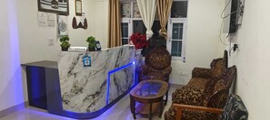Bancada de recepção com tampos de mármore, corrimão iluminado em azul e uma placa com a inscrição 'RECEPTION'. Duas sofás com padrão e uma mesa redonda pequena na área de estar.
