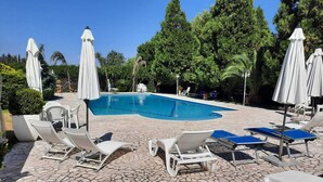 Una piscina con sillones de descanso y sombrillas, rodeada de árboles y un patio de piedra.