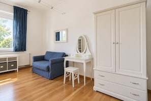 Une pièce avec un canapé bleu, une coiffeuse blanche avec un miroir, une armoire blanche et une fenêtre avec des rideaux.