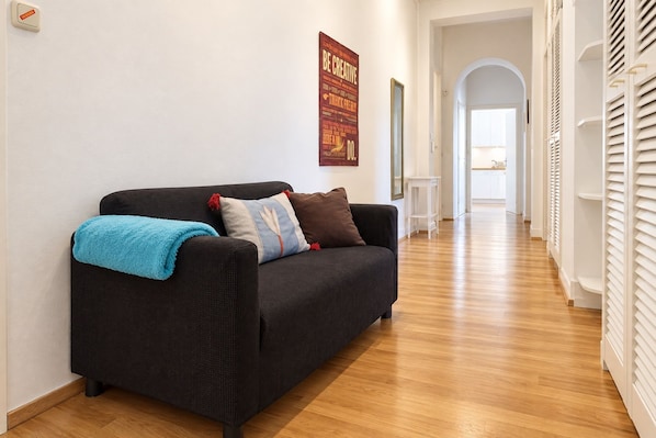 Un salon moderne avec un canapé foncé, un plaid bleu et des coussins décoratifs. Une affiche avec un texte motivant est accrochée au mur, et un couloir mène à une cuisine.