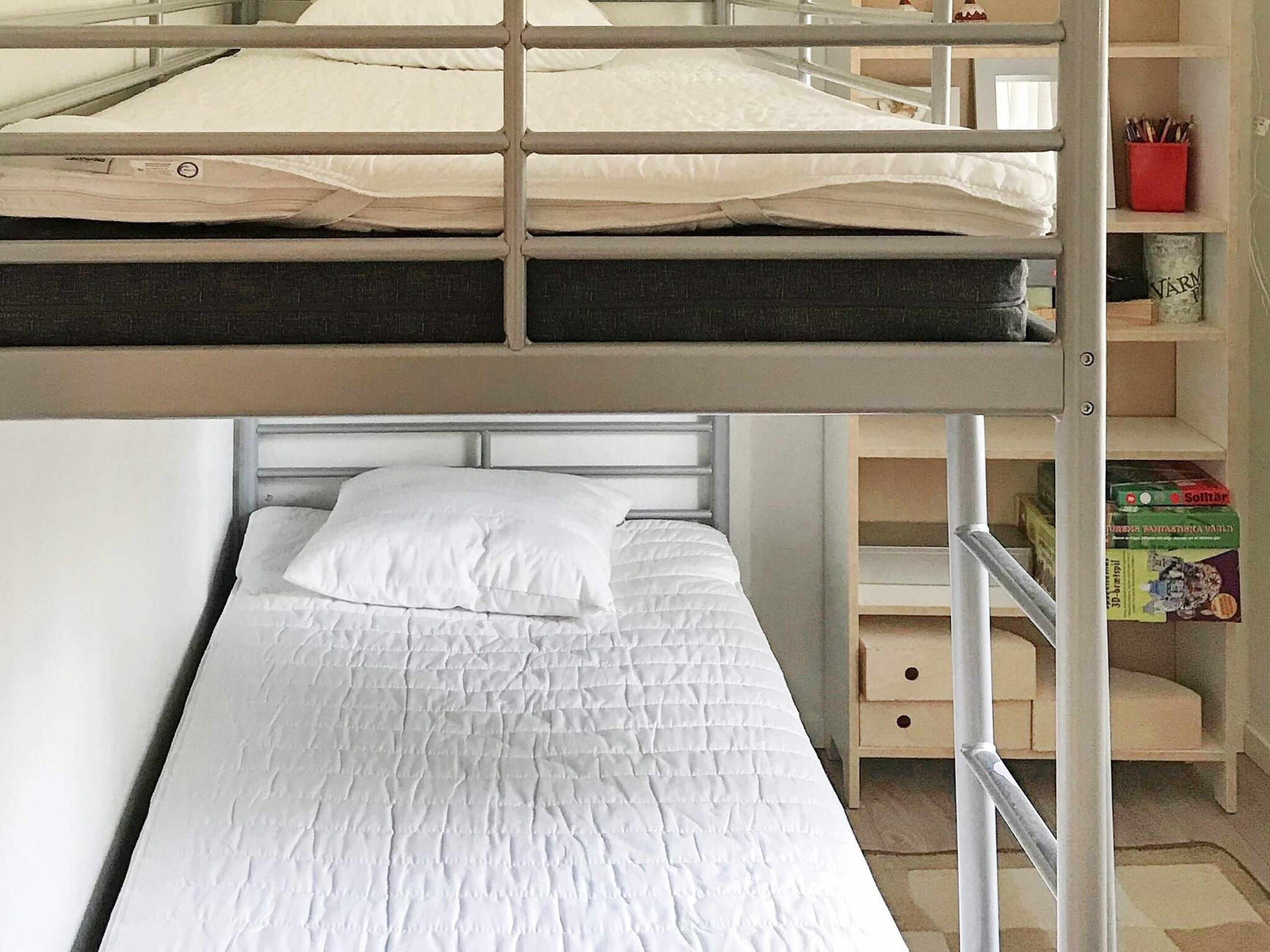 Una cama litera con una cama inferior y una cama individual debajo. Hay un pequeño estante de madera con un recipiente rojo y algunos libros.