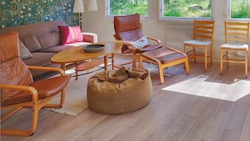 A living room with a sofa, chairs, a coffee table, and a bean bag. There are windows with a view of greenery outside.