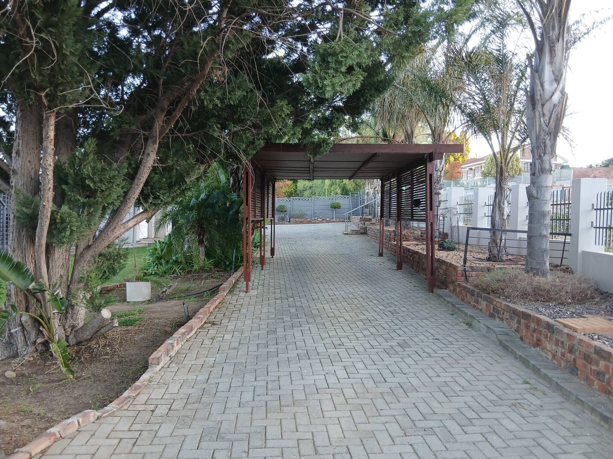 A paved pathway leading to a covered area with a pergola, surrounded by trees and a brick wall.
