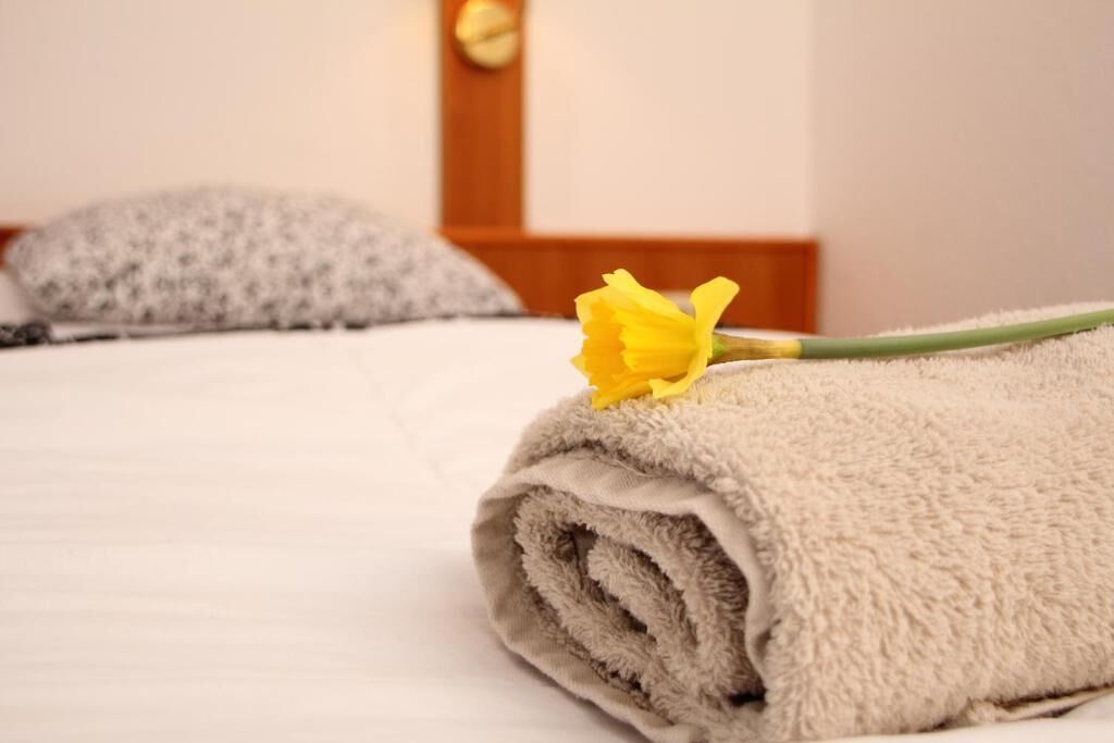 A neatly made bed with a rolled towel and a single yellow flower.