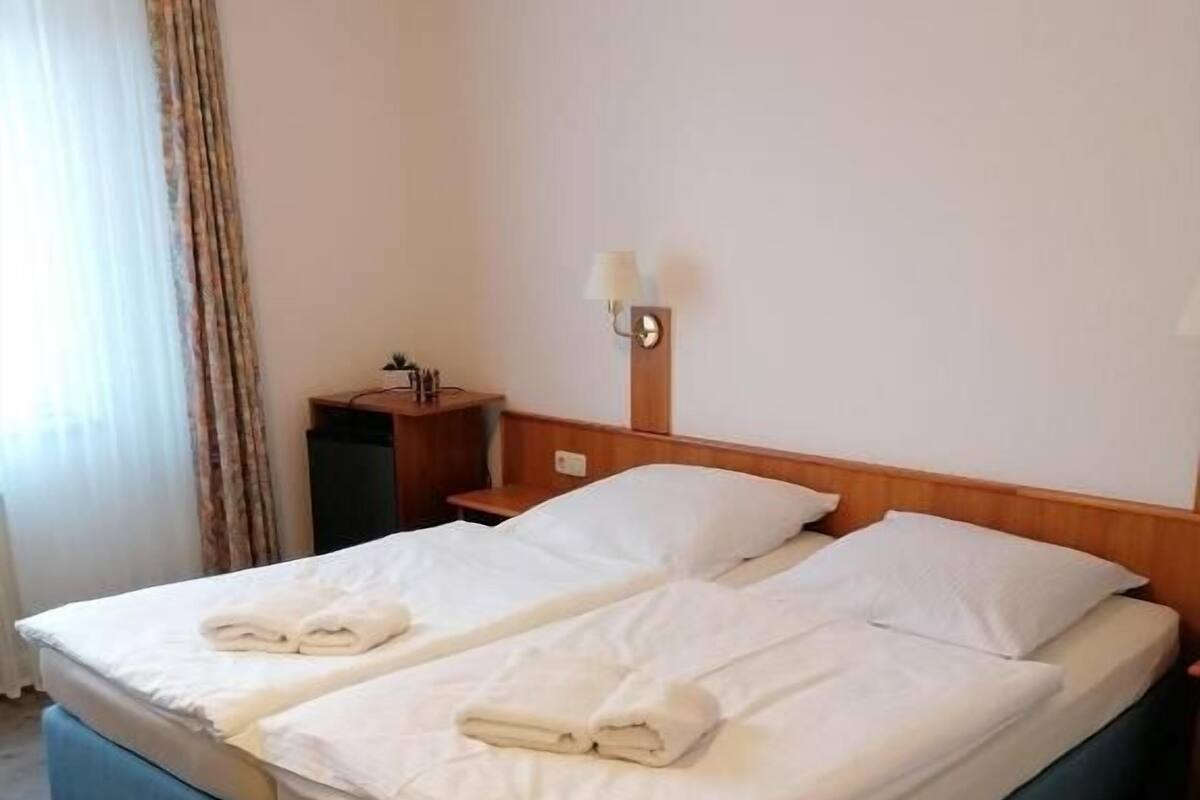 A double bed with white linens and towels, a bedside table with a lamp, and a wooden nightstand.