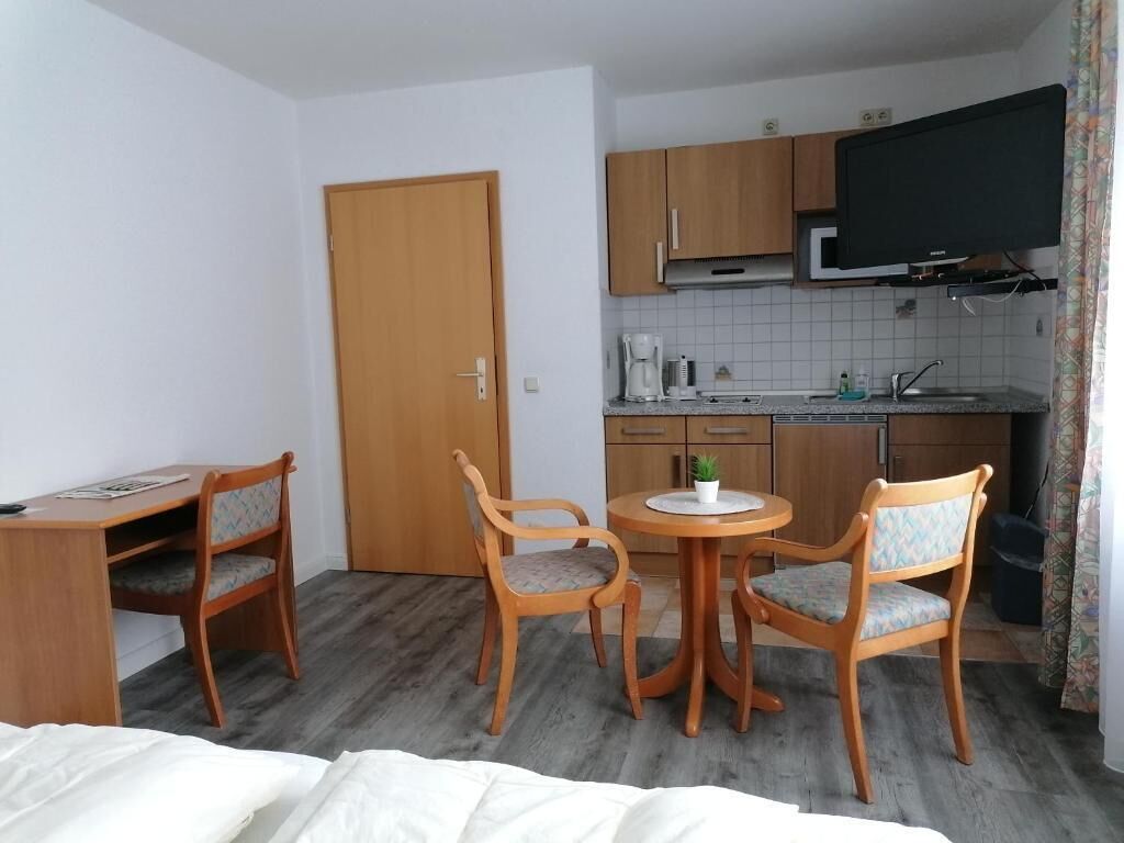 Ein gemütliches Hotelzimmer mit Bett, Schreibtisch, zwei Stühlen, einem kleinen runden Tisch, einem Fernseher und einer Küchenzeile.