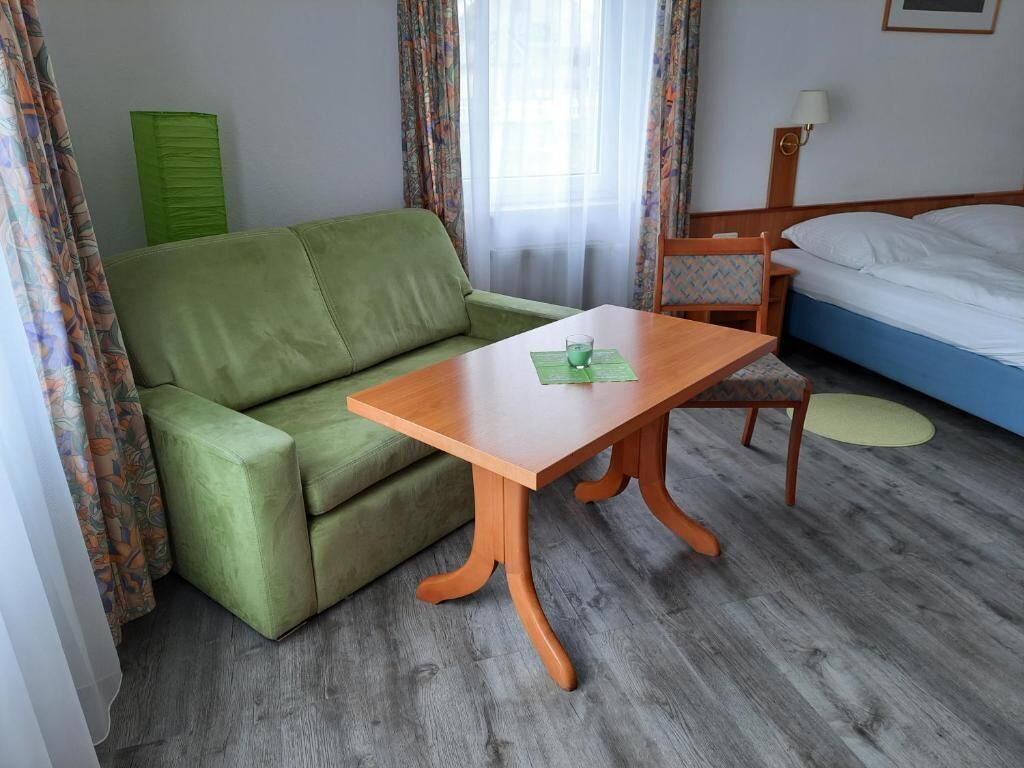 Una stanza con un divano verde, un tavolo in legno, un letto con biancheria bianca e un poggiatesta verde, oltre a una lampada a parete.