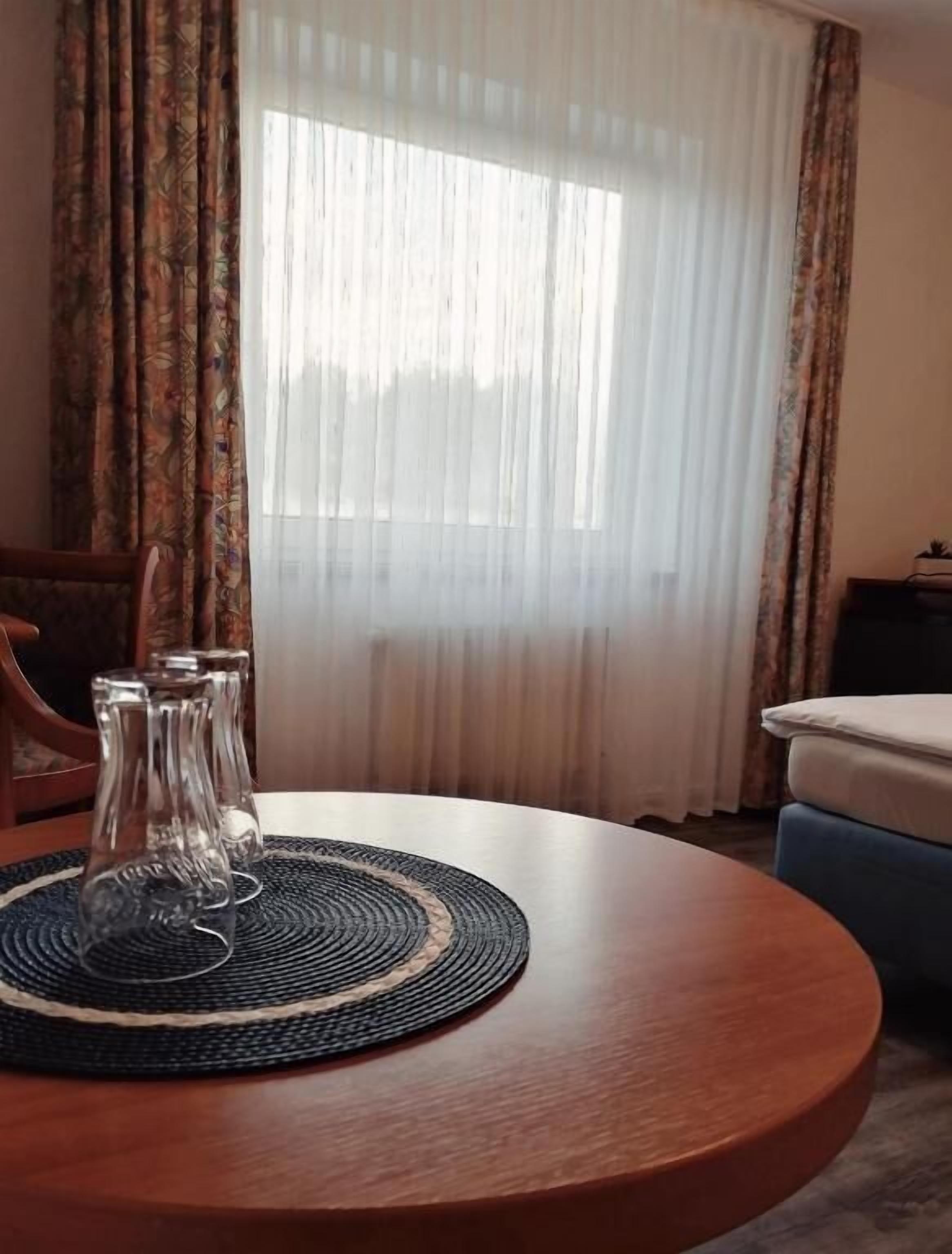 A round wooden table with a woven placemat, two clear glass vases, and a blue sofa in a room with patterned curtains and a large window.