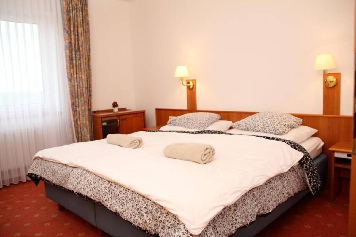 A hotel room with a large bed, two bedside lamps, a nightstand, and a window with curtains.