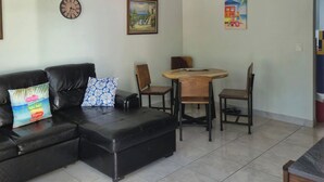 Ein Wohnzimmer mit einer schwarzen Ledersofa, einem Holzesstisch mit Stühlen, einer Uhr und zwei gerahmten Kunstwerken an den Wänden.