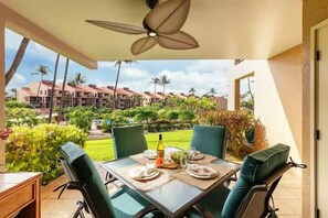 A dining area with a table set for four, a ceiling fan, and a view of a resort complex. - Maui Bliss Garden Lanai, Dual AC, Beach, Pool, Gym (Kihei)