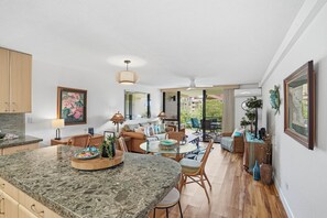 A modern kitchen with granite countertops, a dining area with a glass table, and a living space with a sofa and chairs. - Maui Bliss Garden Lanai, Dual AC, Beach, Pool, Gym (Kihei)