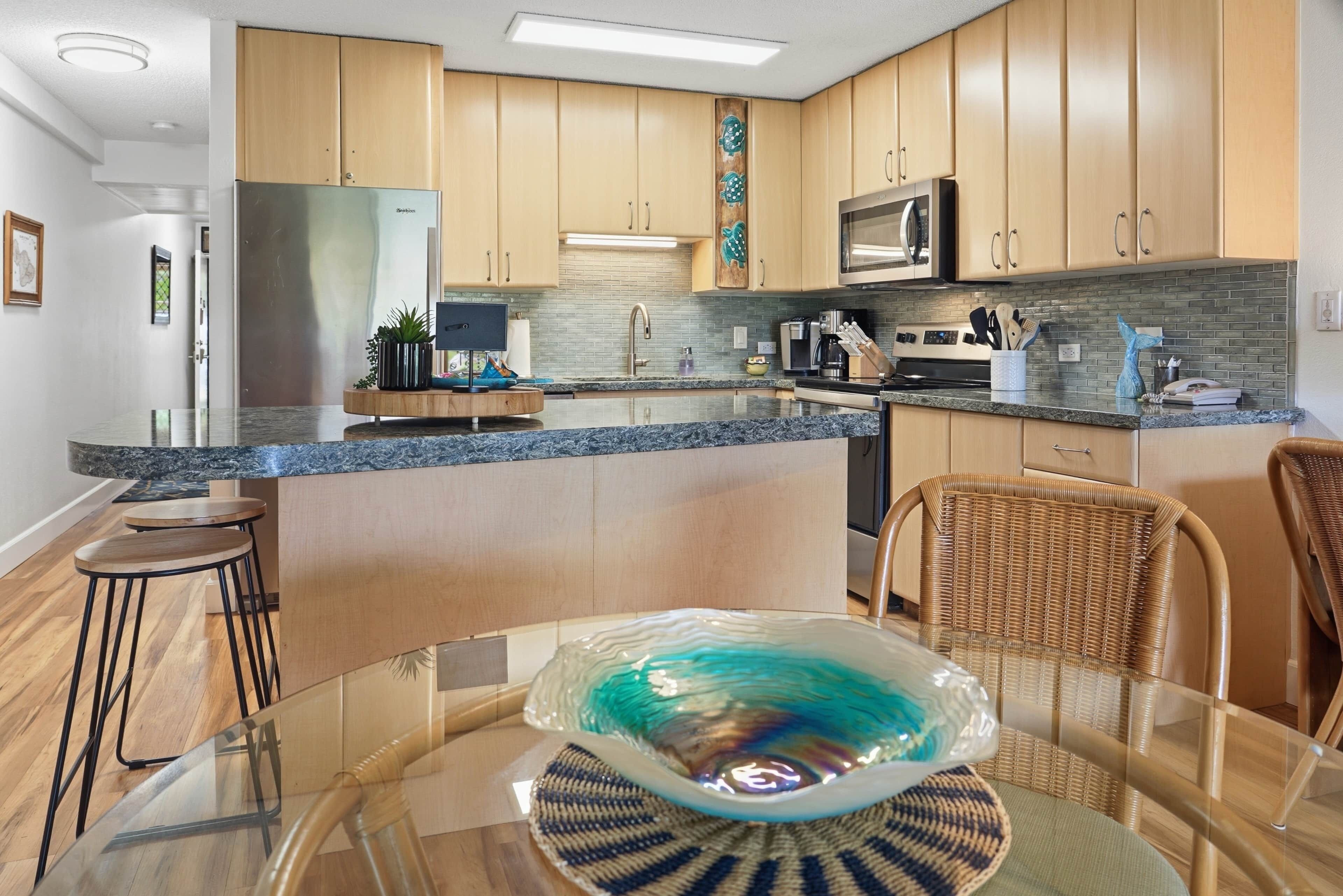 A modern kitchen with wooden cabinets, a granite countertop, a glass dining table, and a wicker chair.