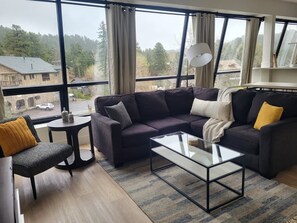 A modern living room with a dark-colored sofa, a glass coffee table, and a view of a street and trees through large windows.