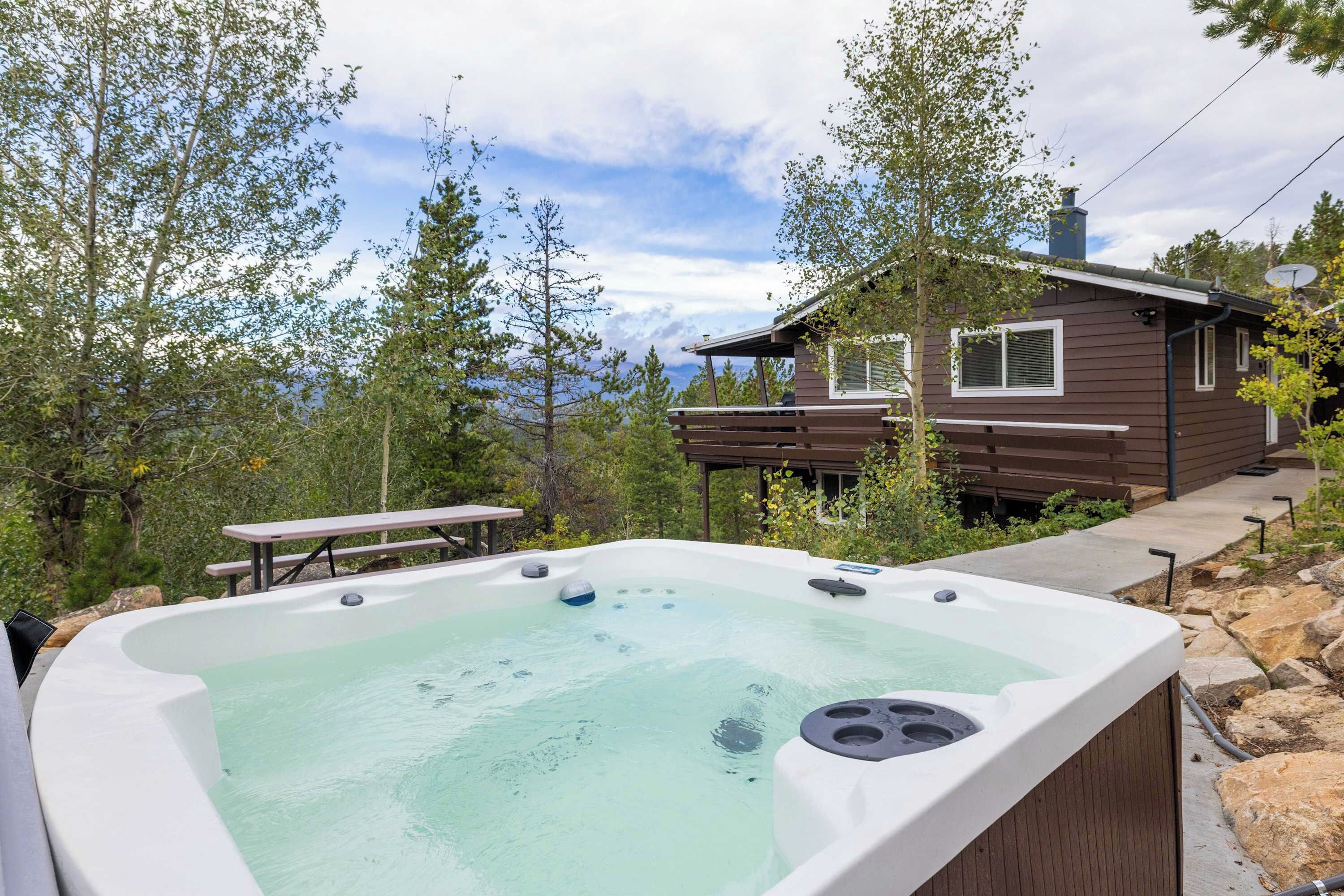 A hot tub with a wooden deck and a picnic table, surrounded by trees and a cabin.