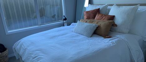 A neatly made bed with white bedding, a bedside lamp, and a window with blinds.