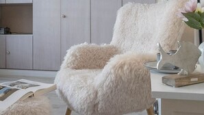 Ein flauschiger weißer Sessel mit Holzbeinen, ein kleiner runder Tisch mit einer Metallskulptur und eine Holzkommode mit Ablage in einem Zimmer mit hellen Wänden und einem Fenster.