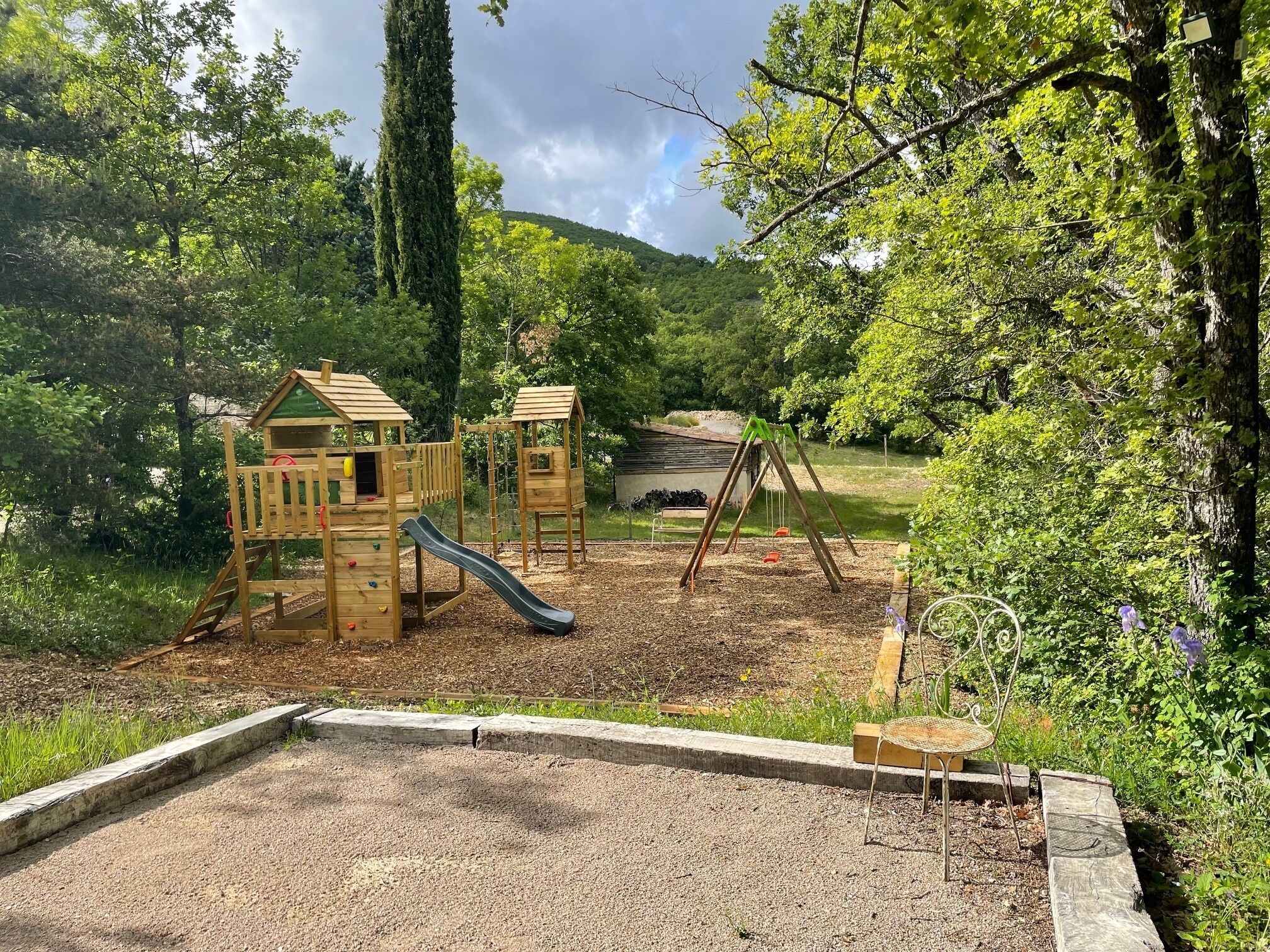 Une aire de jeux dotée de structures en bois, notamment d’un toboggan et d’échelles d’escalade, entourée d’arbres et d’une zone recouverte de gravier.