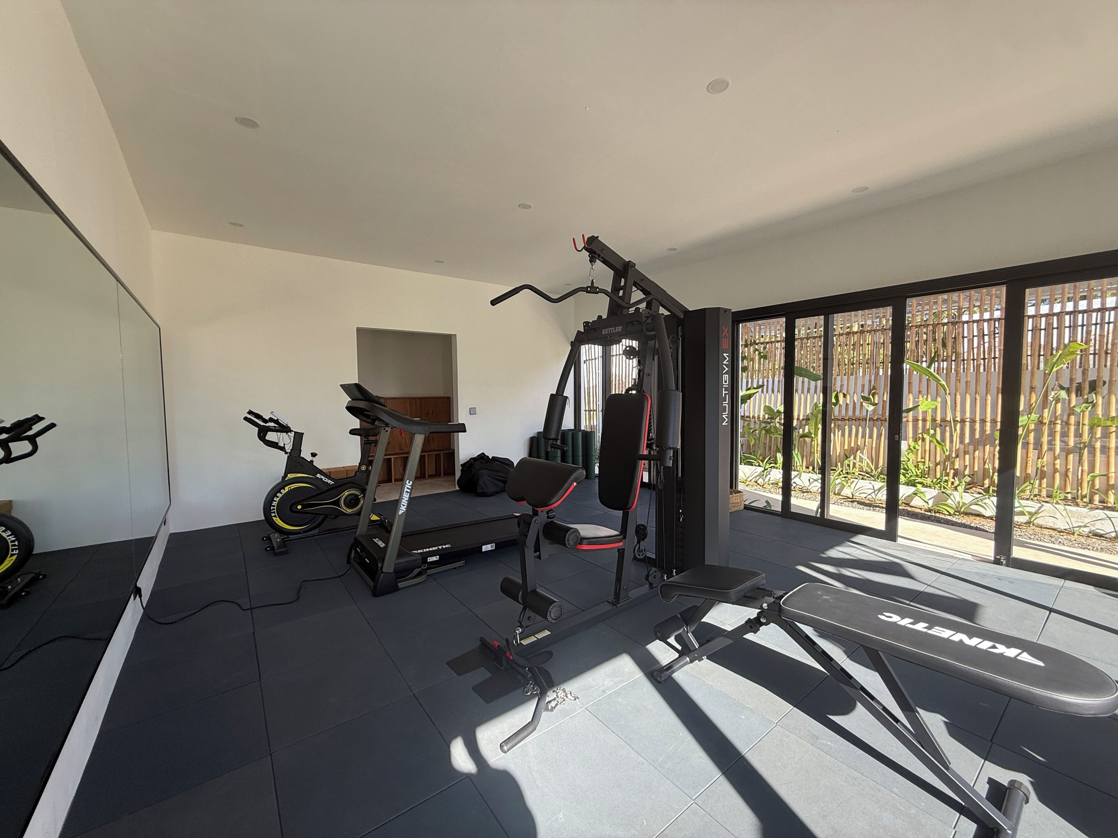 Un gimnasio en casa con equipo de ejercicio, una cinta de correr y un amplio ventanal con vistas a un jardín.