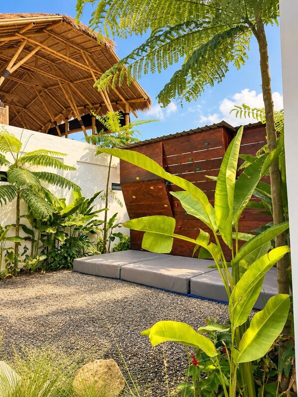 A covered outdoor seating area with a thatched roof, surrounded by lush greenery and a wooden fence.