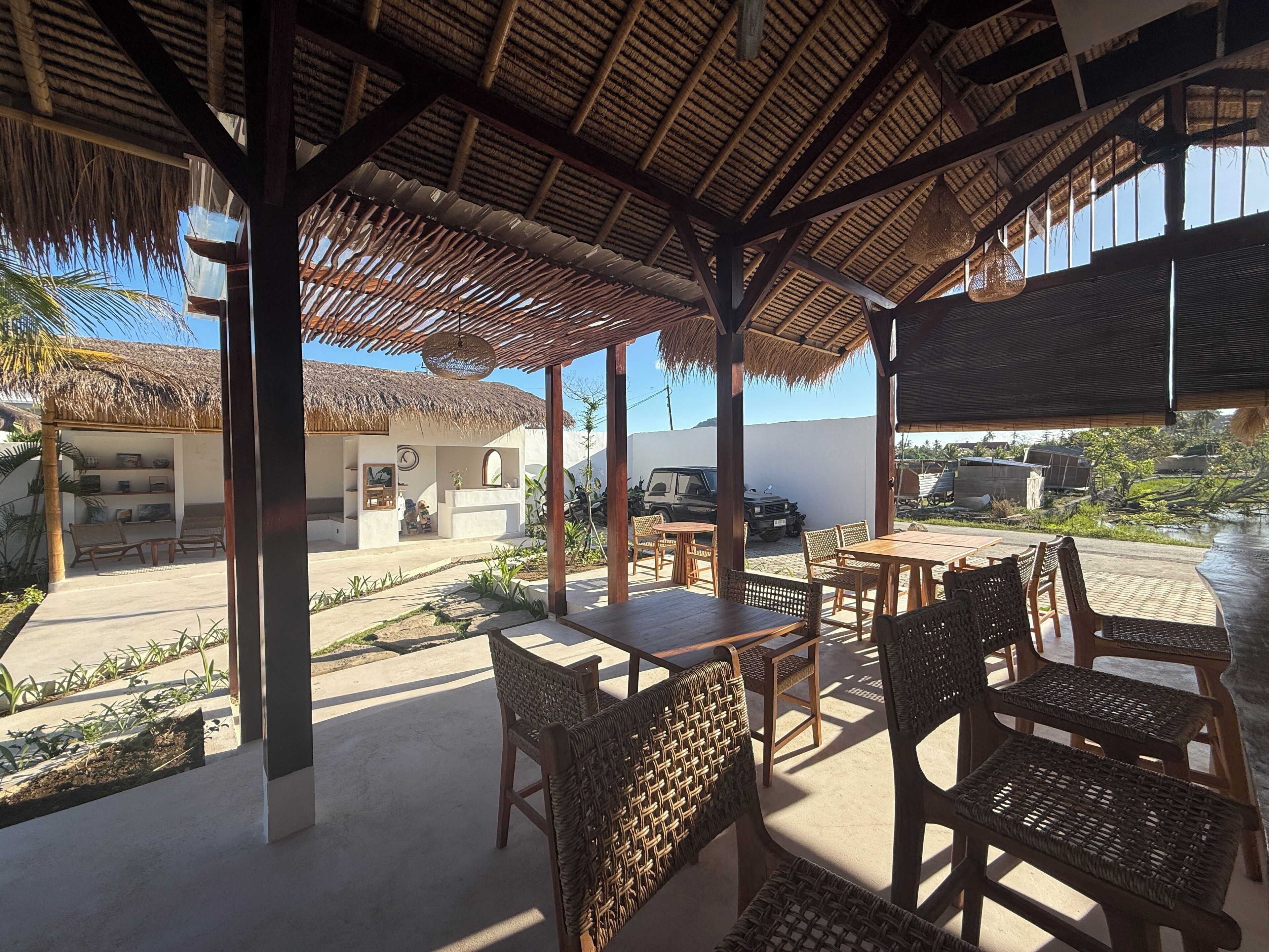 A covered outdoor seating area with tables and wicker chairs, a thatched roof, and a view of a building and vehicles in the background.