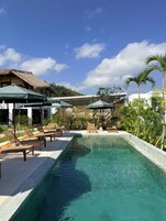 Zona de piscina con sillas de descanso y sombrillas, rodeada de plantas tropicales y un telón de fondo de montañas.