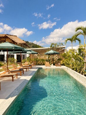 A swimming pool with lounge chairs and umbrellas, surrounded by tropical plants and a wooden fence. - Kairos boutique sports hotel (Selong Belanak)