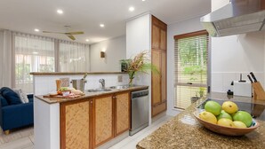 Una cocina moderna con lavavajillas incorporado, un armario de madera, una encimera con un cuenco de fruta y una ventana con persianas.