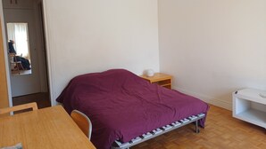 Ett sovrum med en säng, ett träbord, en stol, ett litet träbord som används som nattduksbord och en vit hylla.