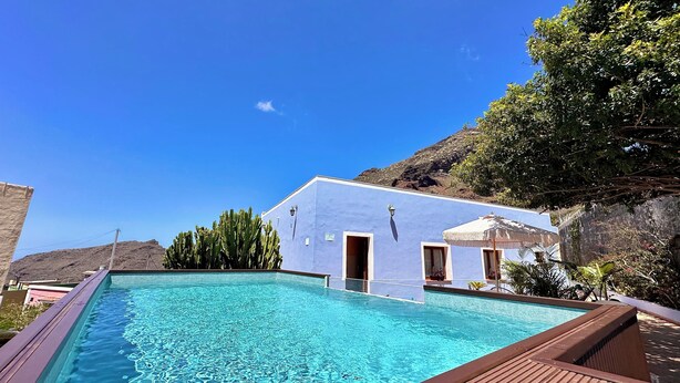 Un'area piscina con un edificio blu, un solarium in legno e uno sfondo montuoso.