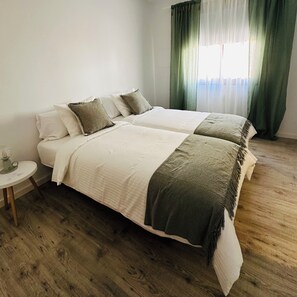 A double bed with a white and green bedspread, a small bedside table, and a window with green curtains.