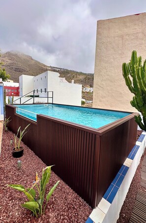 A rooftop swimming pool with a wooden deck, surrounded by plants and a view of a mountain.