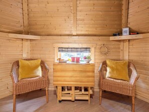 A wooden cabin interior with two wicker chairs, a wooden table, a window with a view, and a shelf with items.
