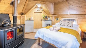 Quarto acolhedor de cabana de madeira com lareira, cama com almofadas, pequena cozinha e teto de madeira.