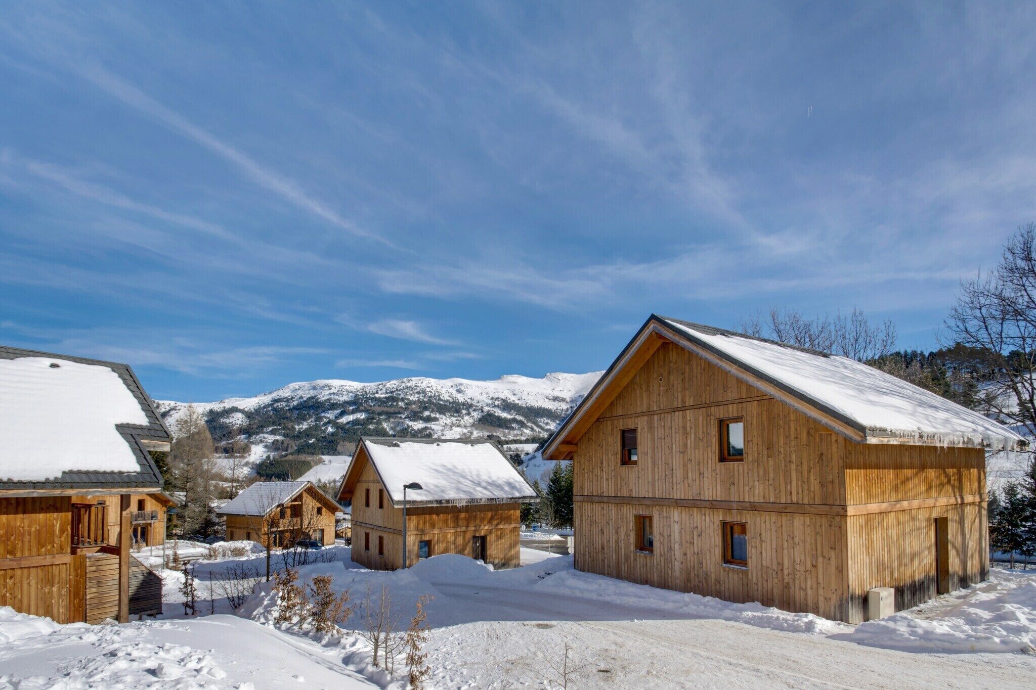 Eine Holzhütte mit schneebedecktem Dach, umgeben von ähnlichen Hütten, vor einer Kulisse schneebedeckter Berge.