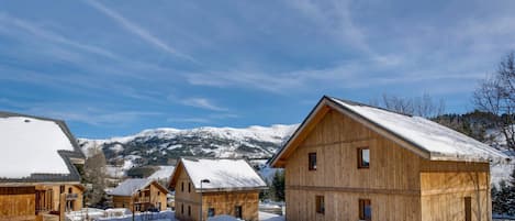 Eine Holzhütte mit schneebedecktem Dach, umgeben von ähnlichen Hütten, vor einer Kulisse schneebedeckter Berge.