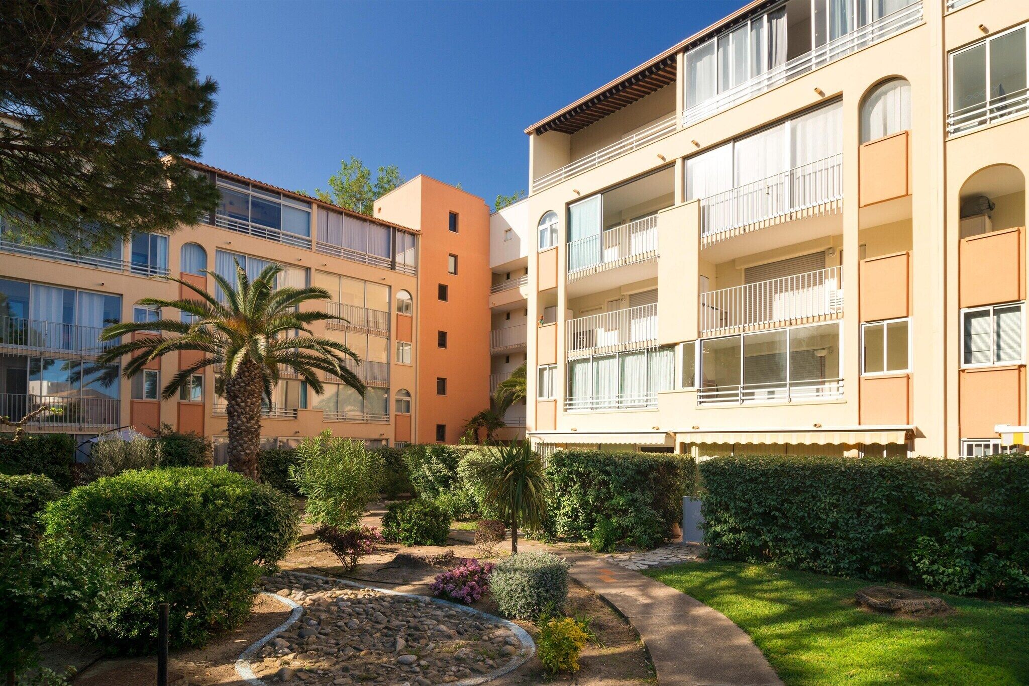 A multi-story building with balconies, a garden with various plants, and a clear sky.