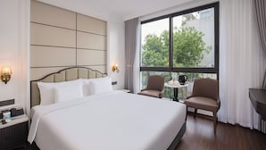 Habitación de hotel con una cama grande, dos sillones, una mesita y vistas a árboles a través de un ventanal amplio.