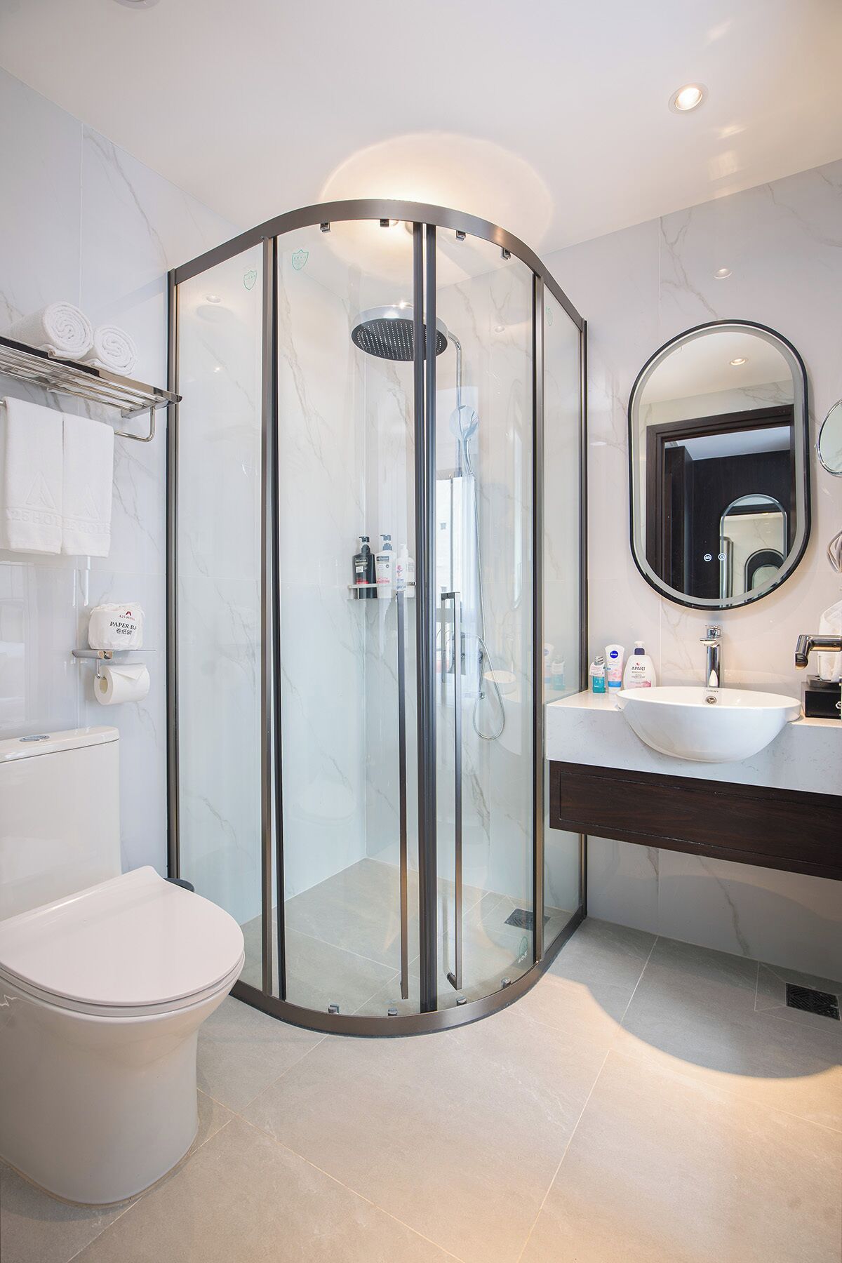 A modern bathroom with a glass-enclosed shower, a toilet, a sink with a mirror, and toiletries on the counter.