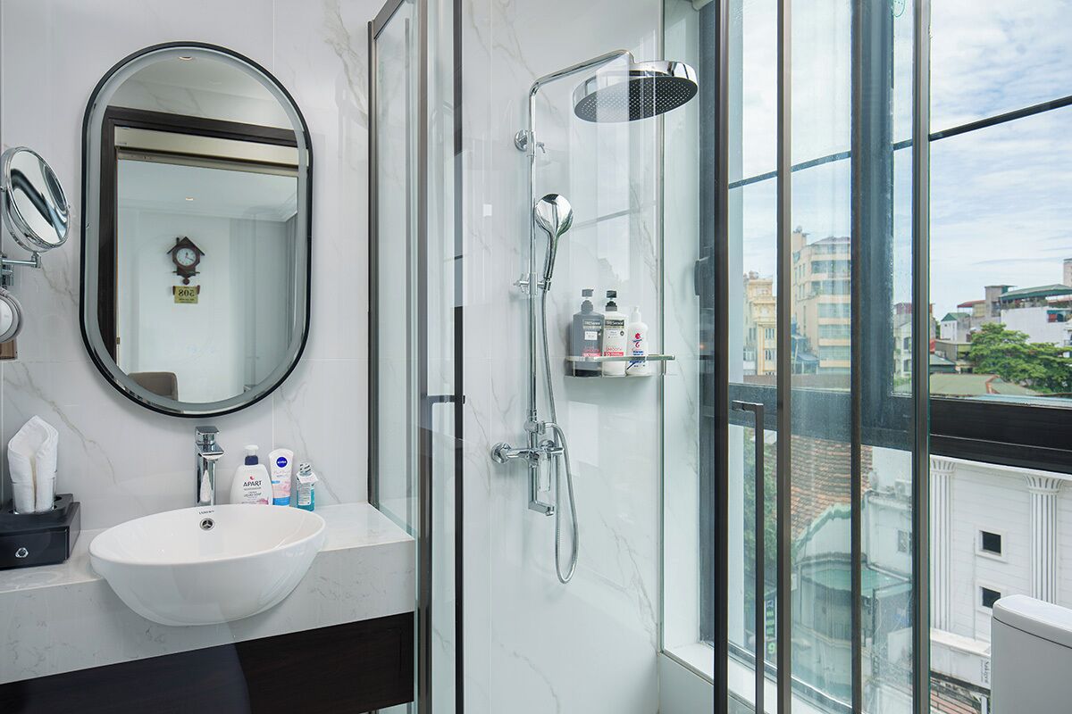A modern bathroom with a large mirror, a white sink, a glass-enclosed shower, and a view of the city through large windows.