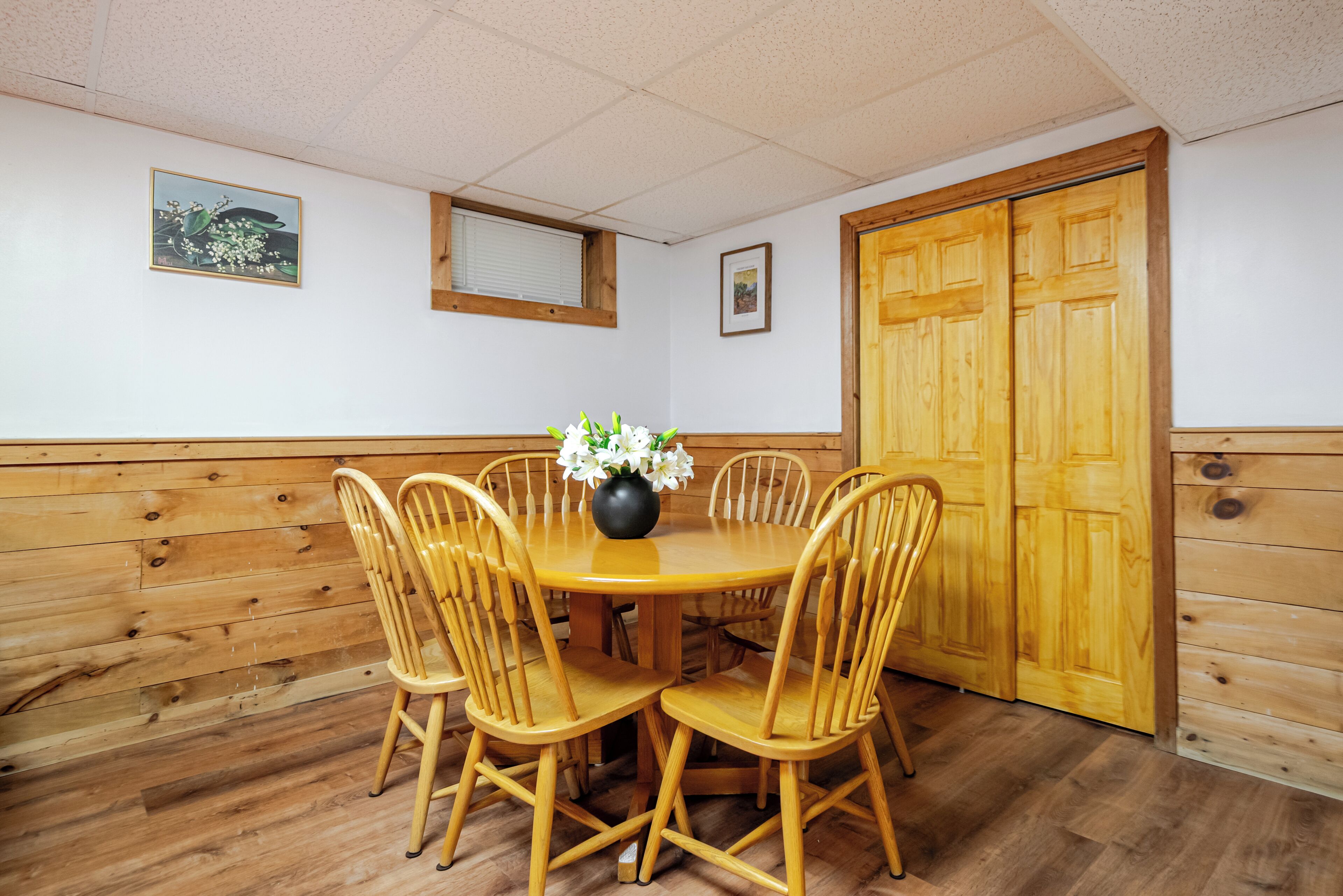 A dining area with a round wooden table, four wooden chairs, a vase with flowers, and a wooden door.