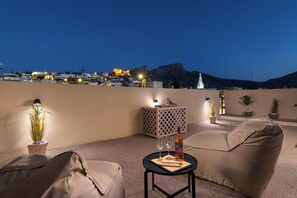 A rooftop terrace with a sofa, a small table with a bottle and glasses, potted plants, and a city view at dusk. - Antheia Traditional House in Archaggelos (Rhodes)