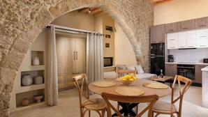 Um arco de pedra que dá acesso a uma área de refeições com uma mesa redonda, cadeiras e uma cozinha ao fundo.