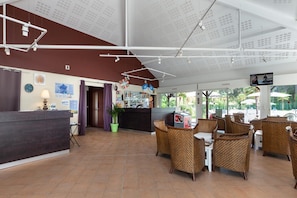 Un hall d’hôtel avec un comptoir d’accueil, des meubles en osier, une horloge et une grande fenêtre donnant sur l’extérieur.