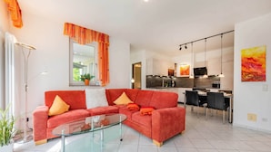 Uma sala de estar moderna com uma mesa de centro de vidro, um sofá vermelho com almofadas laranja, uma área de jantar com mesa e cadeiras pretas, e uma cozinha ao fundo.