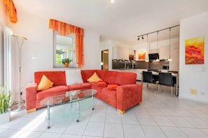 A modern living room with a glass coffee table, a red sofa with orange cushions, a dining area with a black table and chairs, and a kitchen in the background. - House Dünenwald 15 - Sunny Island: 57 m², terrace & 300 m to the sandy beach (Zinnowitz)