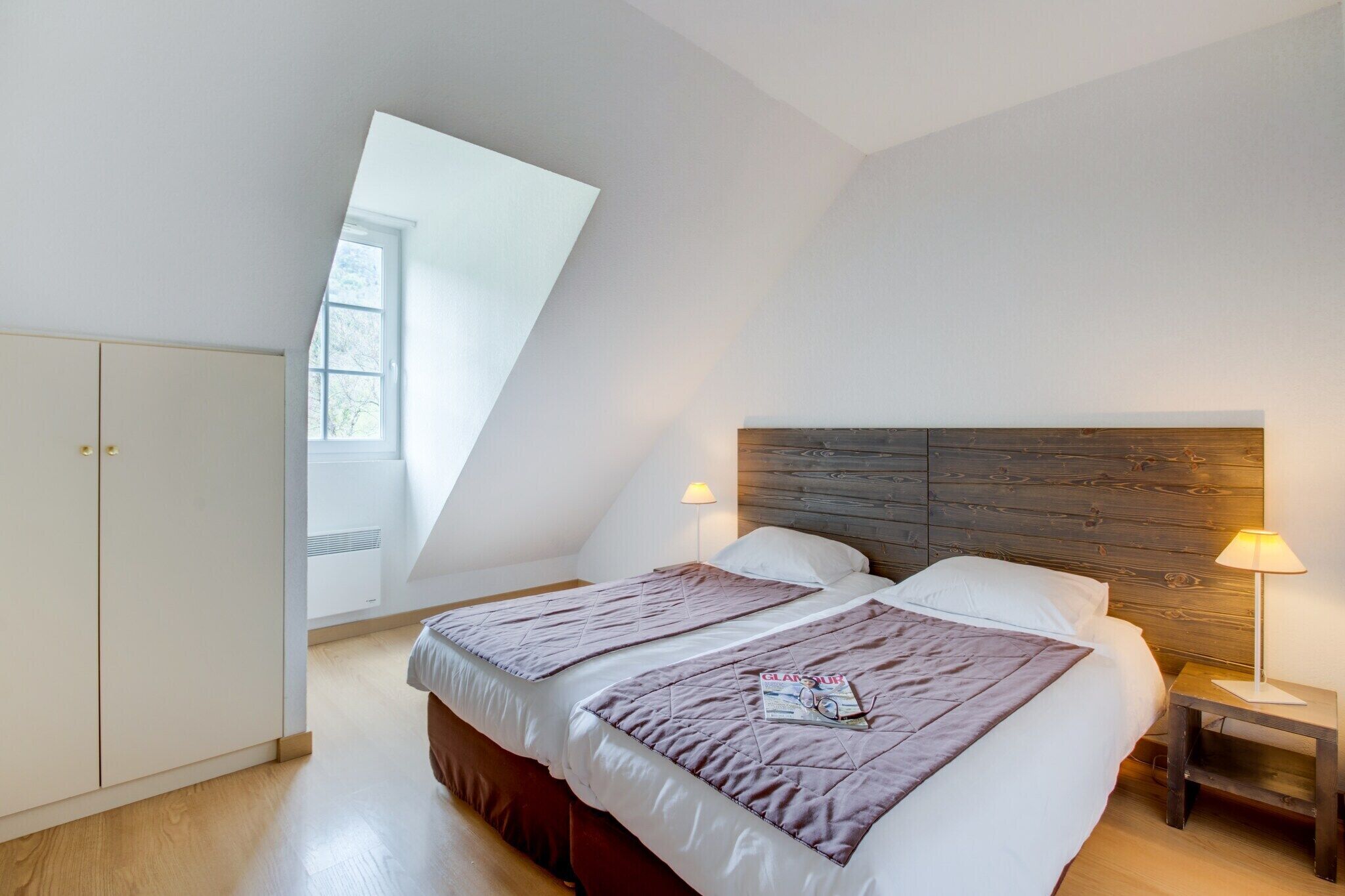 Un dormitorio con dos camas, cabecero de madera, lámparas de noche y una ventana.