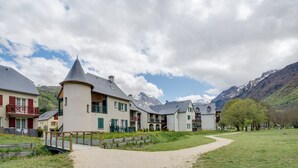 Een rij traditionele Europese gebouwen met een centrale toren, een geplaveid pad en bergen op de achtergrond.